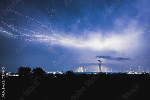 Long, branched, powerful lightning bolts strike down behind the trees. Dramatic lightning bolt. High quality photo