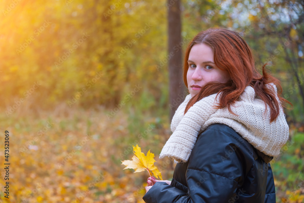 portrait of a red - haired smiling girl in a jacket and scarf with maple leaves in her hand . against the background of autumn nature, the concept of human emotion