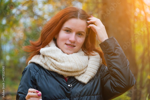 portrait of a red-haired smiling girl in a jacket and scarf straightening her hair against the background of autumn nature and the bright sun the concept of human emotion