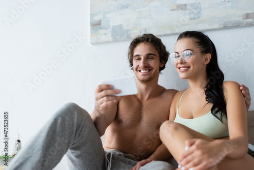 Smiling shirtless man holding blurred smartphone near smiling girlfriend in eyeglasses in bedroom