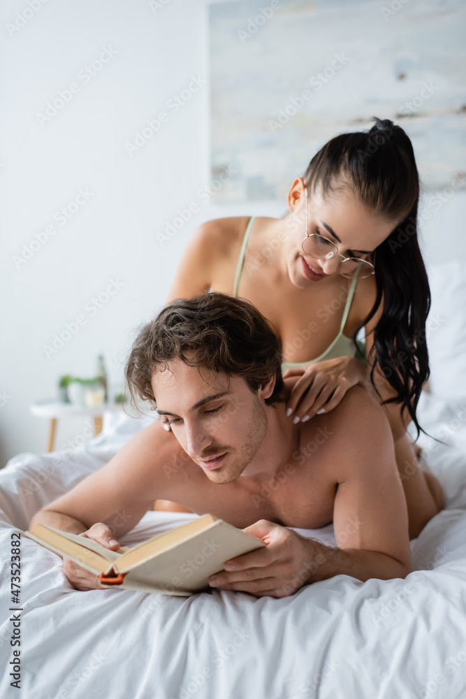 Smiling woman in eyeglasses touching shirtless man with book on bed Smiling woman in eyeglasses touching shirtless man with book on bed
