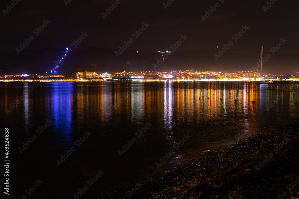 Night city with lights and reflections in the water and mountains near the sea and red sky, pebble shore.