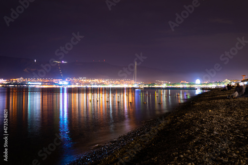 Night city with lights and mountains by the sea and red sky, pebble shore.