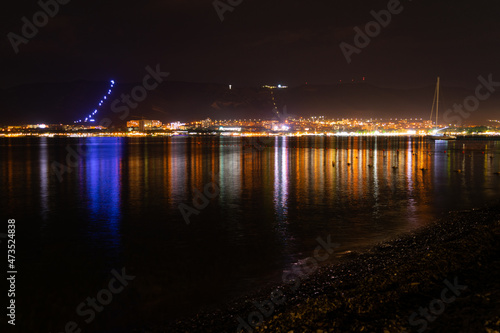 Night city with lights and reflections in the water and mountains near the sea and red sky, pebble shore.
