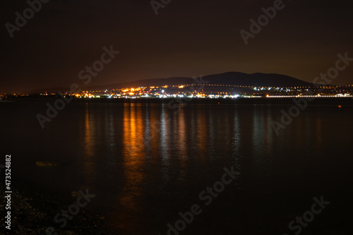 Night city with lights and reflections in the water and mountains by the sea and red sky, pebble shore, very beautiful at night.