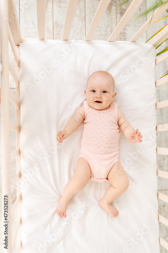 smiling baby girl in a crib in a pink bodysuit six months on a white cotton bed laughing, top view