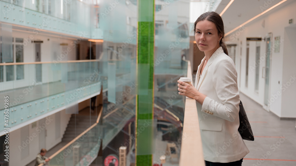 Fototapeta premium young business woman in a white jacket is standing in a public space with a takeaway coffee in her hand. Confident brunette freelancer, portrait on the background of a business center.