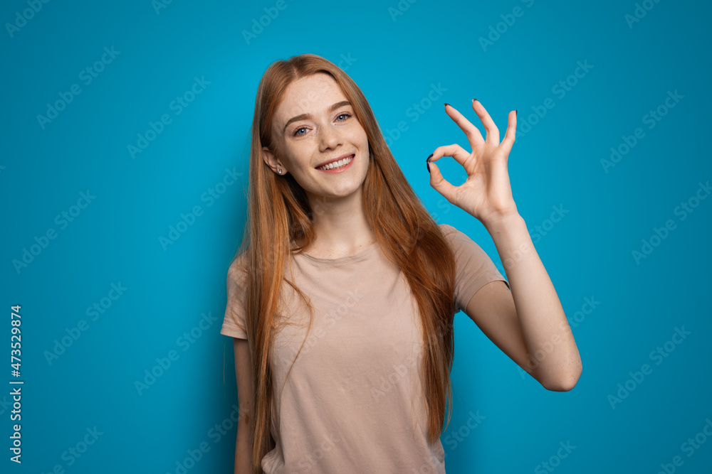 Portrait of smiling redhead woman looking at camera, showing okay sign standing over blue background. No problem gesture. Portrait of smiling redhead woman looking at camera, showing okay sign standing over blue background. No problem gesture.