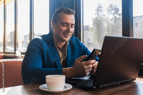 A business happy man stylish smiling businessman in an attractive European-looking suit works in a laptop, listens to music and drinks coffee sitting at a table in a cafe by the window