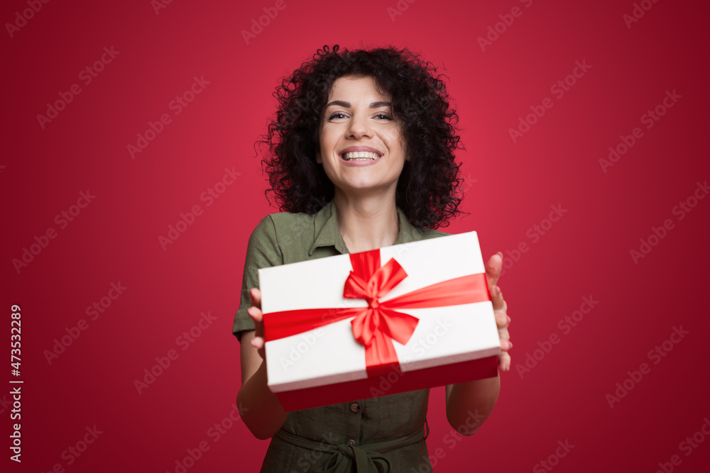 Close up photo of a curly woman giving box anniversary to you isolated on red background. Smiling Caucasian woman. Close up photo of a curly woman giving box anniversary to you isolated on red background. Smiling Caucasian woman.