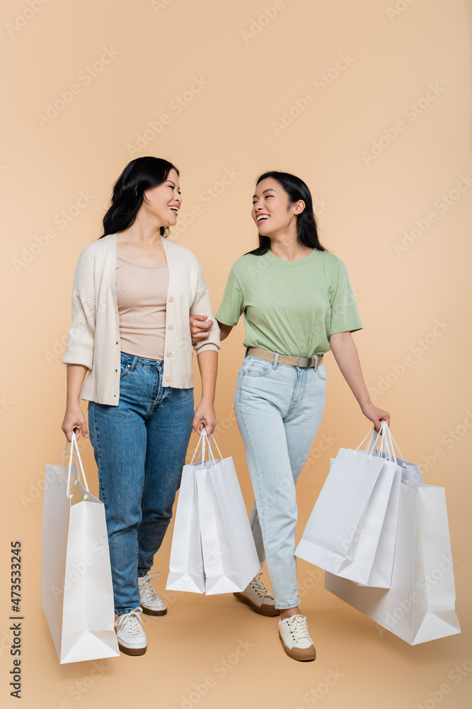 full length of asian mother and happy young adult daughter smiling while holding shopping bags on beige full length of asian mother and happy young adult daughter smiling while holding shopping bags on beige