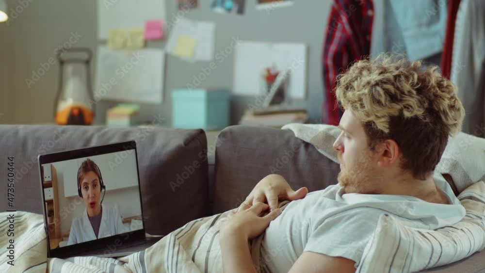 Sick man lying on sofa at home and speaking with female doctor on video call on laptop during online healthcare consultation Sick man lying on sofa at home and speaking with female doctor on video call on laptop during online healthcare consultation