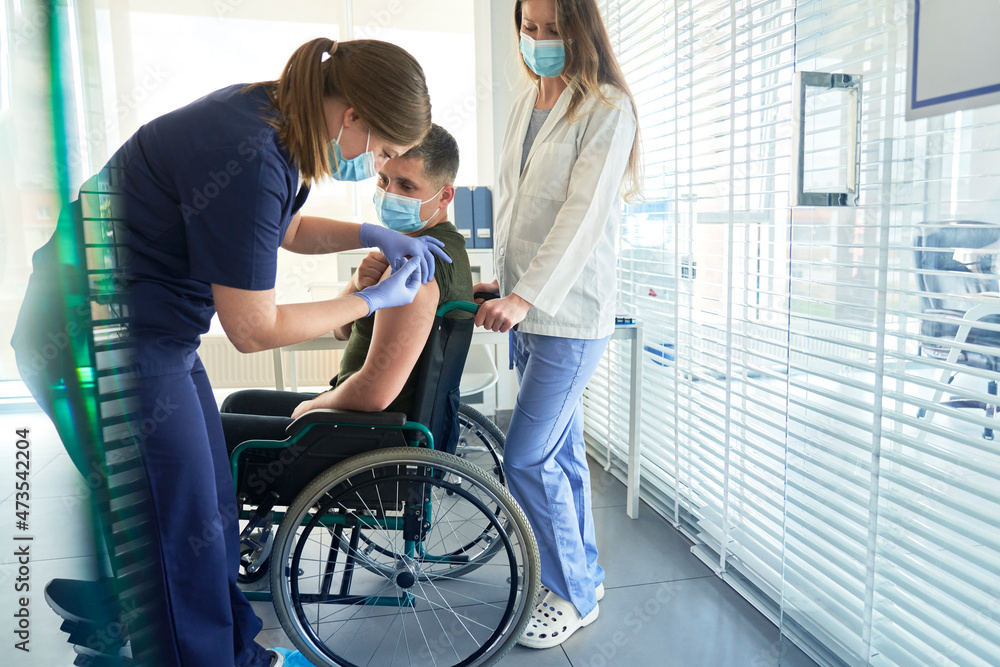Female doctor giving COVID-19 vaccination dose to disabled man sitting on wheelchair at clinic Female doctor giving COVID-19 vaccination dose to disabled man sitting on wheelchair at clinic