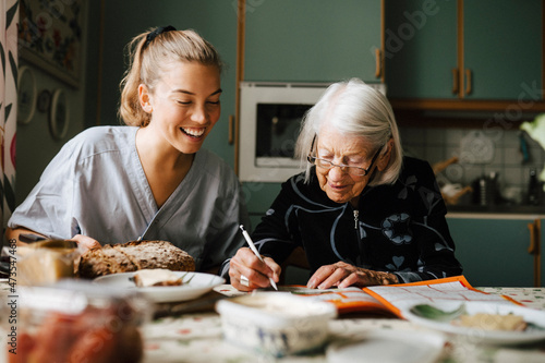 Senior woman solving crossword puzzle in book sitting by smiling nurse in kitchen at home photo