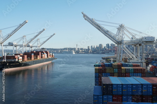 Container SSA terminal with gantry cranes on both sides. There is a container merchant vessel being loaded and discharged on the left. Skyline of Seattle on the horizon. 
