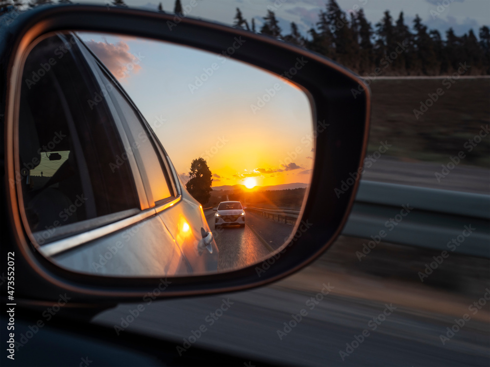 The sunset is reflected in the mirror of a car racing down the highway