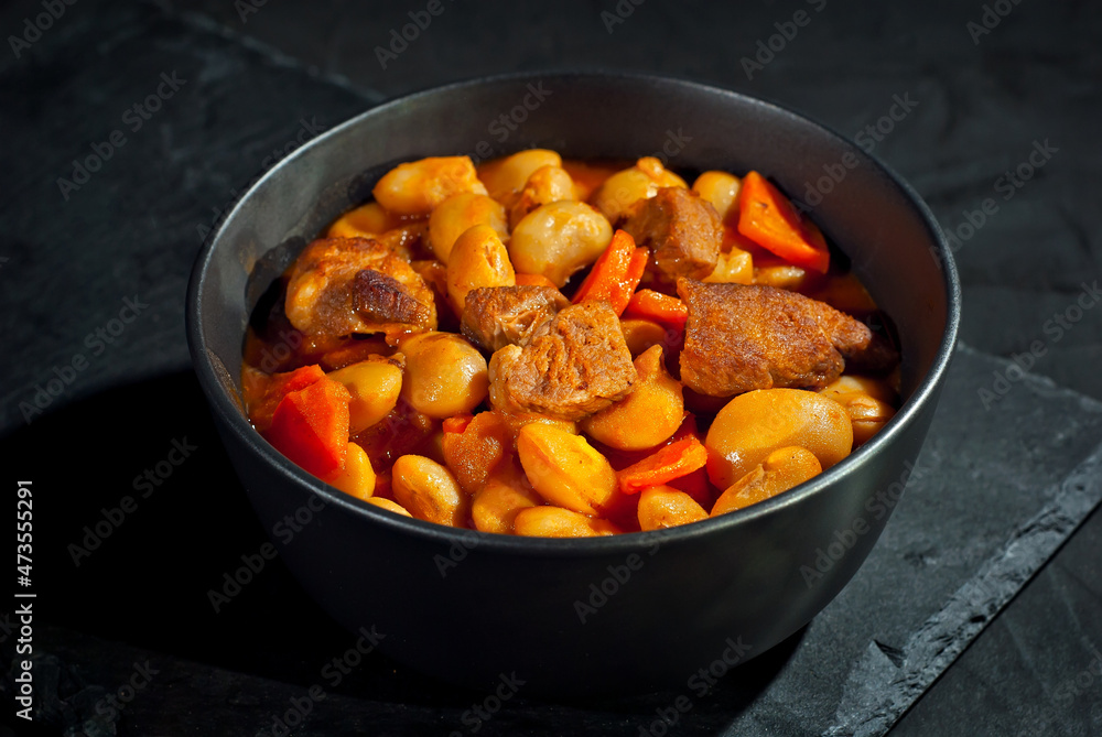Yellow beans in a black bowl. Boiled beans with meat on a stone table. Beans with tomato sauce and pork. Contrasting dramatic light as an artistic effect. Yellow beans in a black bowl. Boiled beans with meat on a stone table. Beans with tomato sauce and pork. Contrasting dramatic light as an artistic effect.