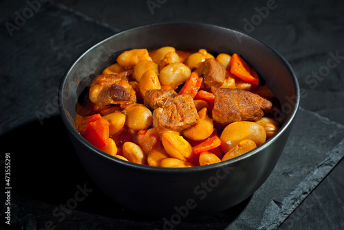 Yellow beans in a black bowl. Boiled beans with meat on a stone table. Beans with tomato sauce and pork. Contrasting dramatic light as an artistic effect.
