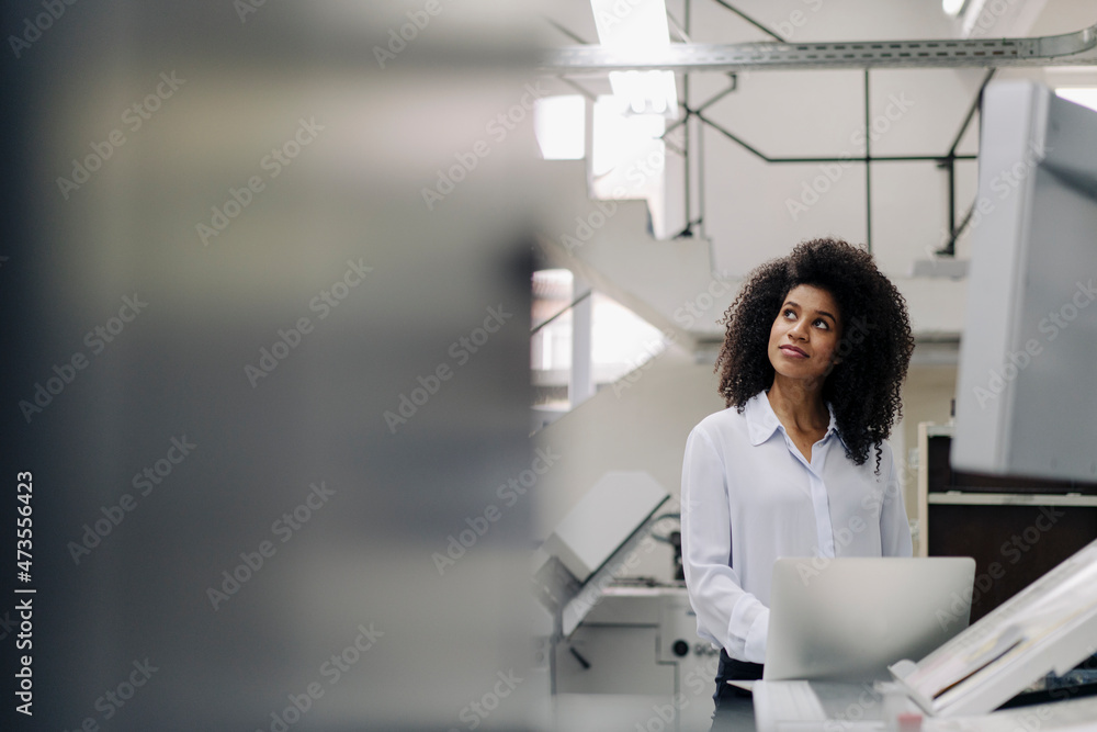 Businesswoman with laptop looking away while standing at machinery in industry Businesswoman with laptop looking away while standing at machinery in industry