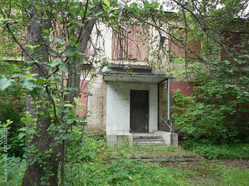 An abandoned two-story brick building with windows covered with metal and boards, with an area overgrown with trees. An abandoned two-story brick building with windows covered with metal and boards, with an area overgrown with trees.