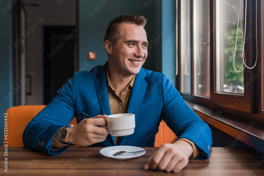 A stylish, positive businessman of Caucasian appearance, a man in a jacket and shirt sits at a table in a cafe on a coffee break and looks away or out the window A stylish, positive businessman of Caucasian appearance, a man in a jacket and shirt sits at a table in a cafe on a coffee break and looks away or out the window