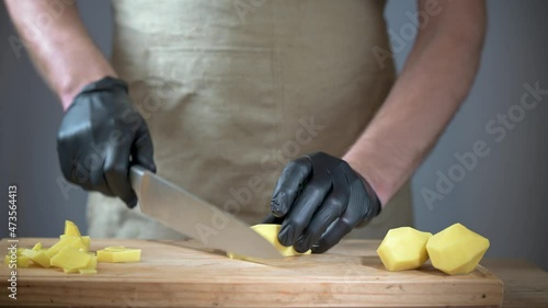 Close-up of men's hands in black disposable gloves. The cook is slicing potatoes on a cutting board. Background in blur.