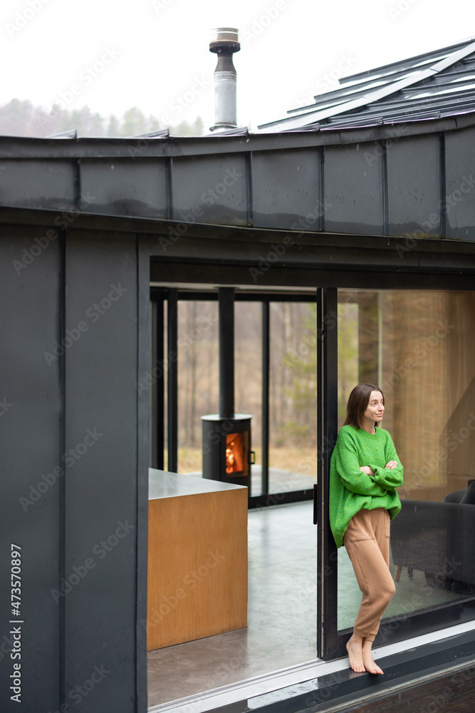Young woman stands at window of a modern house on nature and look away. Concept of comfort and happy modern life. Woman wearing green sweater