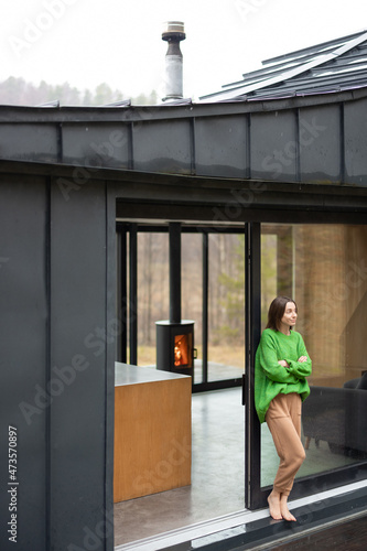 Young woman stands at window of a modern house on nature and look away. Concept of comfort and happy modern life. Woman wearing green sweater