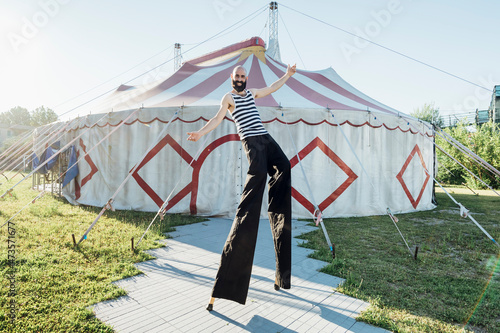 Male artist with arms outstretched walking with stilts on footpath