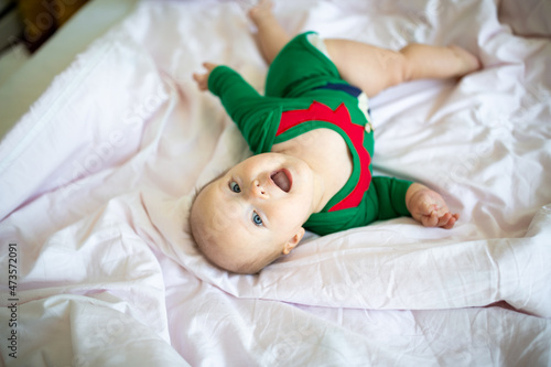 Cute caucasian baby girl six months old wearing elf costume green color. Adorable Santa helper lying on bed at home on white background © Алина Бузунова