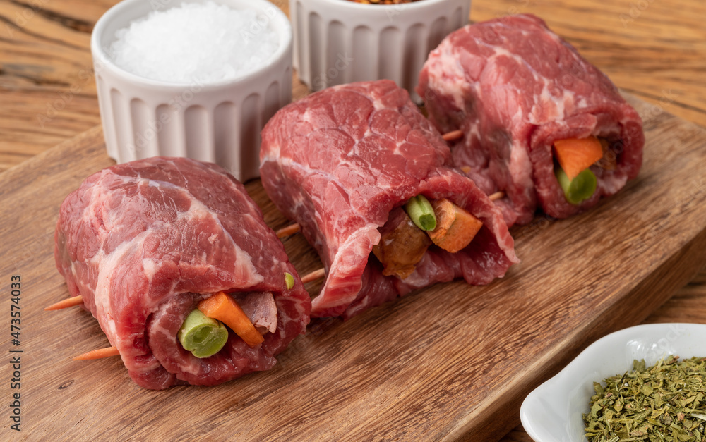 Rolled beef or roulade, vegetable and bacon stuffed meat over wooden table with seasonings Rolled beef or roulade, vegetable and bacon stuffed meat over wooden table with seasonings