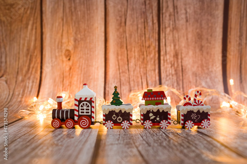 A festive train carries gifts for Christmas. A red toy train. Wooden background with Christmas lights.  Space for text. Christmas card.
