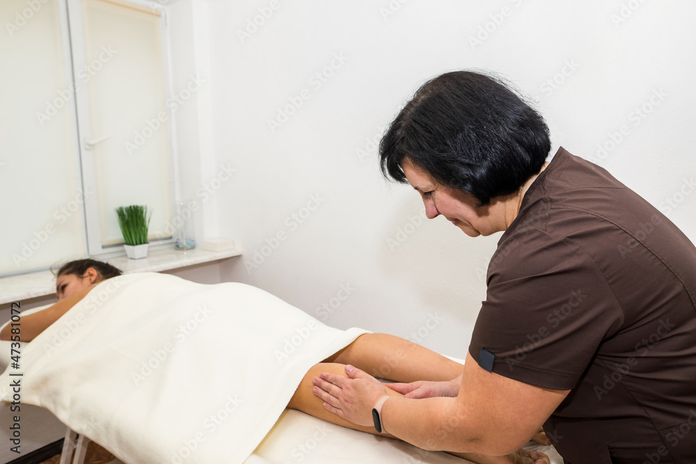 Obraz premium A female massage therapist massages the legs of a young woman lying on a massage table on a white background.