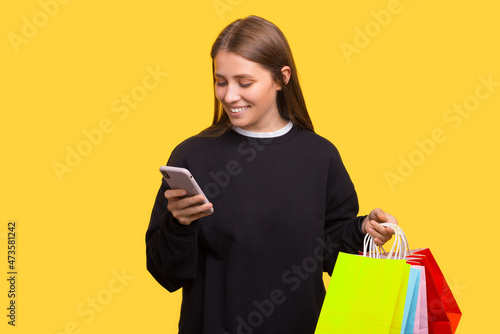 Happy smiling woman with shopping bags is looking at her phone over yellow background.