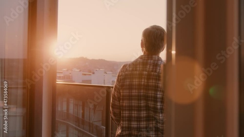 Casual bearded adult man spending morning home, enjoying beautiful senset from balcony and drinking coffee. Cozy apartment. Sceneries. Urban landscape. photo