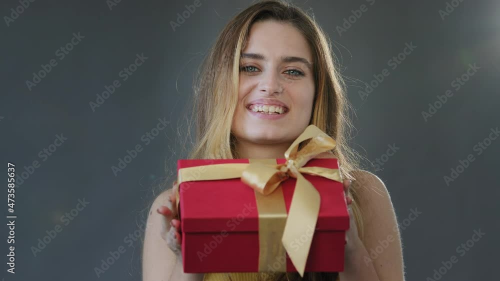 Female studio portrait gray background young beautiful happy generous girl woman holding christmas gift holding red present box with gold ribbon congratulating happy birthday festive mood celebration Female studio portrait gray background young beautiful happy generous girl woman holding christmas gift holding red present box with gold ribbon congratulating happy birthday festive mood celebration