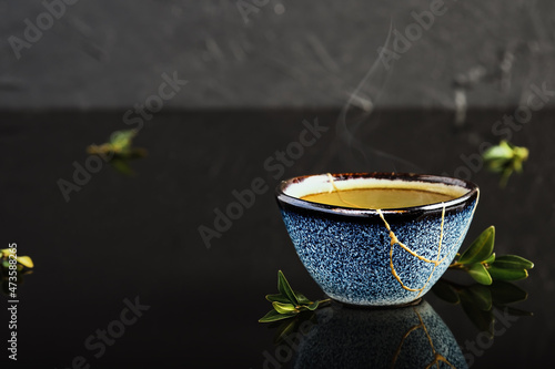Bowl of green Japanese tea, tea leaves lie next the cup. Selective focus on the cup. Steam rises above the bowl. Reclaimed ceramic blue cup, second life of things, recycling or kintsugi photo