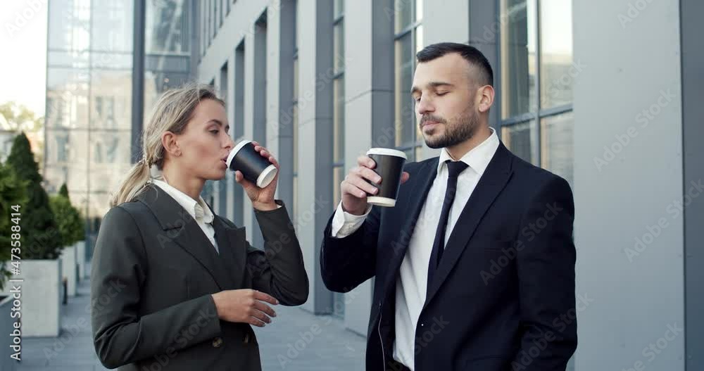 Good looking young male and female business colleagues standing at the street near the big office building, talking to each other and sipping hot coffee to-go Good looking young male and female business colleagues standing at the street near the big office building, talking to each other and sipping hot coffee to-go