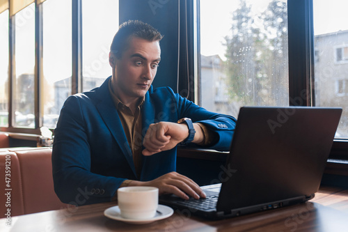 A business surprised man businessman a stylish of Caucasian appearance in a jacket, works in a laptop or computer, sitting at a table by the window in a cafe and looks at the time on a wrist watch