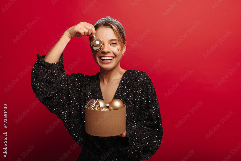 Happy young caucasian girl cheerfully fooling around with New Year's toys on red background. Pretty woman with gathered hair wears shiny black dress. Christmas tree decoration concept Happy young caucasian girl cheerfully fooling around with New Year's toys on red background. Pretty woman with gathered hair wears shiny black dress. Christmas tree decoration concept