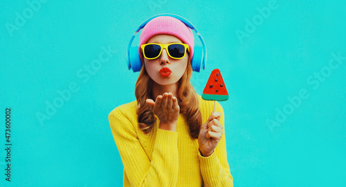 Summer fresh colorful portrait of happy young woman in headphones listening to music with fruit juicy lollipop or ice cream shaped slice of watermelon on blue background