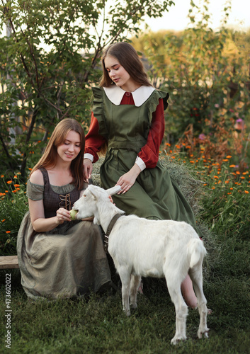 Rural scene. Two beautiful young girls in simple old fashion dresses among trees and flowers playing with goat. Village life with warm atmosphere © Альбина Хусаинова
