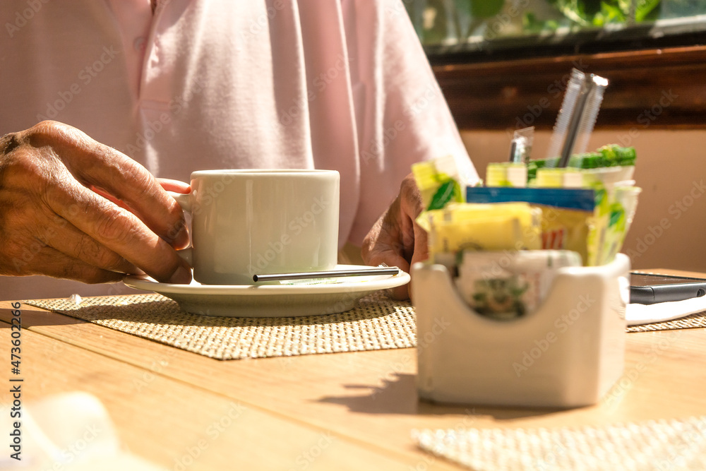 Man's hand holding a white ceramic cup or mug with hot beverage like coffee or tea. Sugar and adulcorants unfocused on foreground. Wooden table, mand with pink shirt