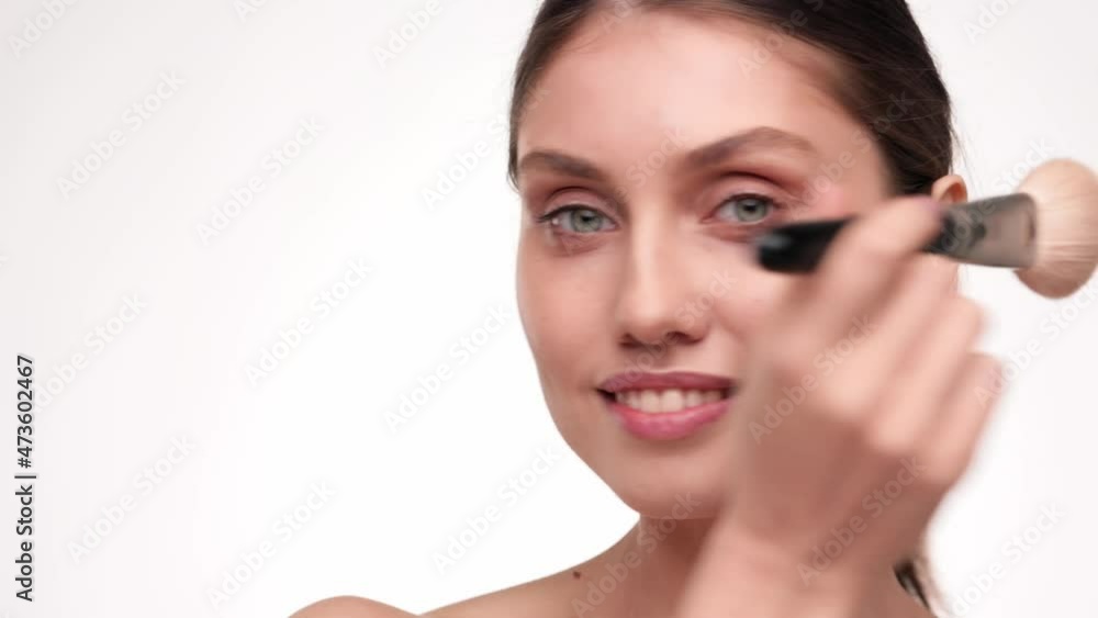 Face pretty female with nude make look at the camera and smiling. Young woman Applying Makeup Blush With Brush. Concept of cosmetology, skincare. Healthy Beauty face on light background at studio. Face pretty female with nude make look at the camera and smiling. Young woman Applying Makeup Blush With Brush. Concept of cosmetology, skincare. Healthy Beauty face on light background at studio.