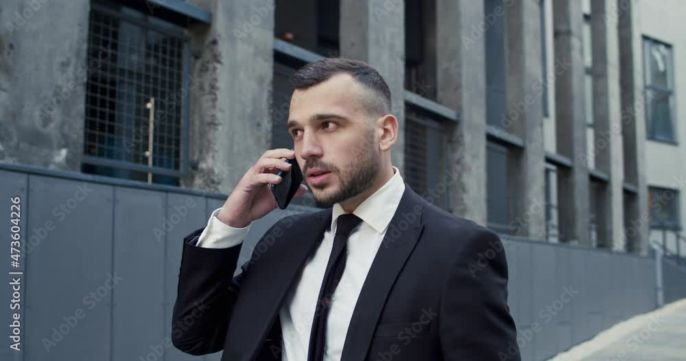 Close up portrait of serious caucasian male professional walking in city and talking on smartphone. Handsome man manager speaking on cellphone on street outdoors. Businessman concept Close up portrait of serious caucasian male professional walking in city and talking on smartphone. Handsome man manager speaking on cellphone on street outdoors. Businessman concept