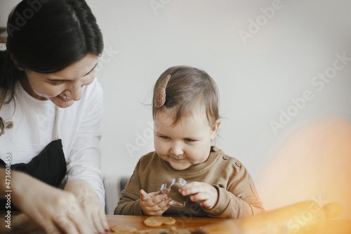 Adorable little daughter with mother making together christmas gingerbread cookies on messy wooden table. Cute toddler girl helps cutting dough with festive cutters for cookies. Family time