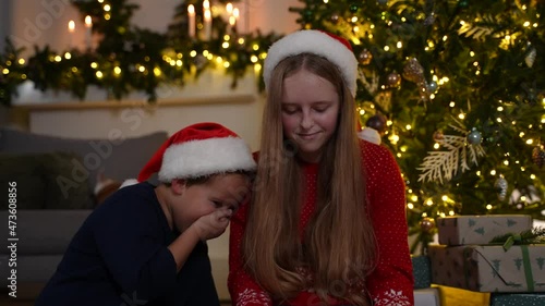 Joyful little brother and his older teenage sister wearing Santa Claus hats sitting on floor near fancy Christmas tree and laughing while watching funny video on tablet, family holiday leisure concept