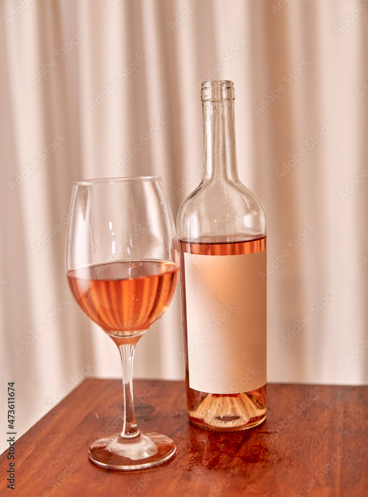bottle of rosé wine with blank label with glass on wooden table with white curtain in the background. vertical. pink wine tasting bottle of rosé wine with blank label with glass on wooden table with white curtain in the background. vertical. pink wine tasting
