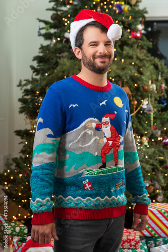 Portrait of a young man with a beard, wearing a Santa Hat and an ugly Christmas sweater standing in front of Christmas tree. photo