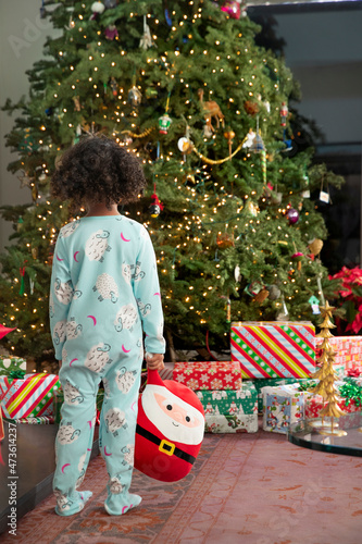 Little girl in onesie pajamas standing and looking at Christmas tree in wonderment.  photo